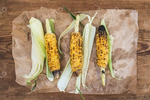 Preview: Grilled corn over oily craft paper and rustic wooden background, top view.