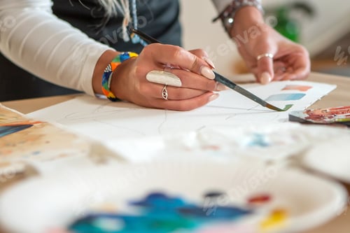 Preview: High angle shot of a person painting with a brush and watercolor paint on a white sheet of paper