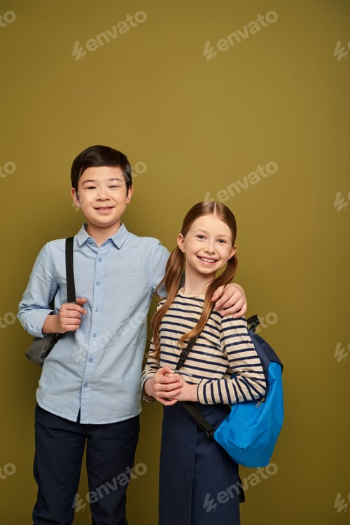 Preview: Smiling asian boy with backpack hugging red haired friend and looking at camera during international