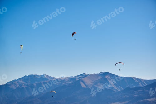 Preview: Drifting easily in the clear sky, paragliders admire the majestic brown mountain peaks.