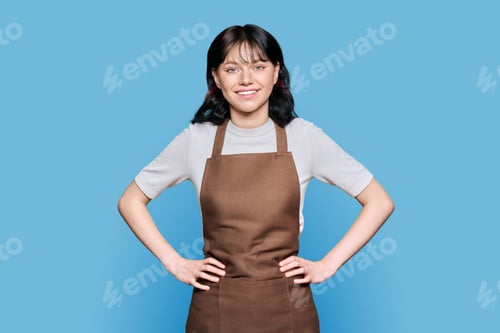 Preview: Smiling young woman staff in apron looking at camera on blue isolated background