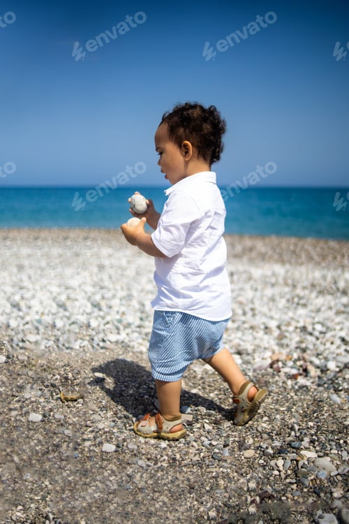 Preview: Curly toddler boy plays on the pebble beach near blue sea