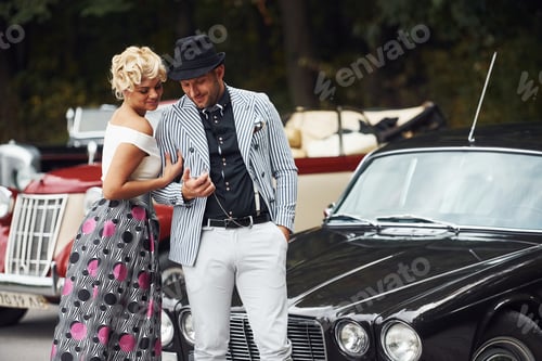 Preview: Beautiful couple in old fashioned wear near retro car with forest at background