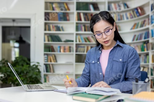 Preview: An asian woman in glasses intently studies in a library, reading and taking notes.