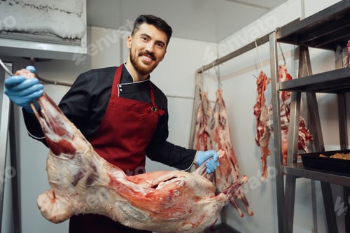 Preview: Male butcher standing by meat hanging on hook in meat shop