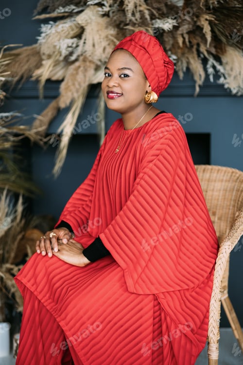 Preview: Pretty African woman wearing an red fashionable suit and headscarf, sitting on the chair in studio