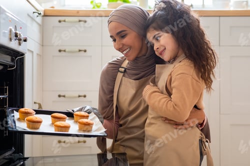 Preview: Happy muslim mother and daughter baking in kitchen, holding tray with muffins
