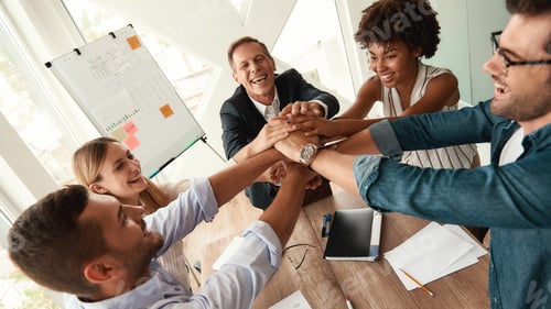 Preview: Happy business people holding hands together while sitting at the office table