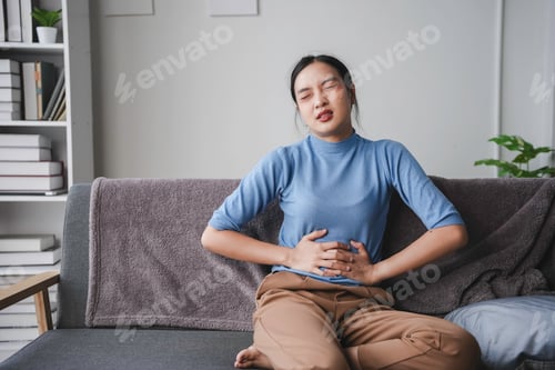 Preview: Young woman is sitting on her sofa, hunched over in pain and holding her stomach