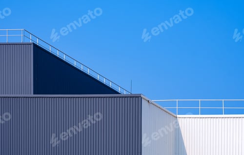 Preview: Gray and white corrugated factory building with steel fence on rooftop against blue sky background