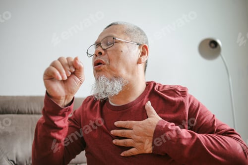 Preview: Senior Sick Man Sitting On Sofa At Home And Holding Hand On Chest While Coughing
