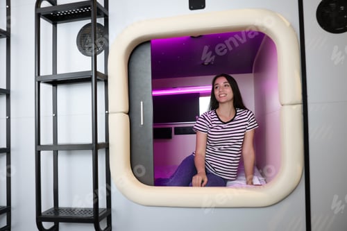 Preview: Happy young woman sitting in capsule of pod hostel