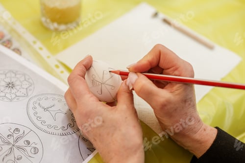 Preview: Easter concept. Adult Hands Drawing on Easter Egg with Pencil.