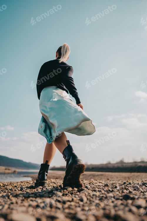 Preview: Woman in silk skirt and brutal trendy boots walking in near river on coast Young lady in rough shoes