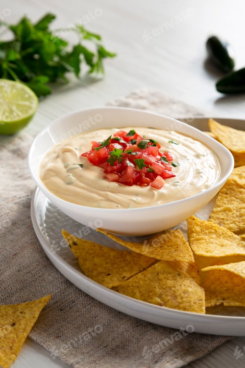 Preview: Homemade cheesy dip in a bowl, yellow tortilla chips over white wooden surface, side view. Close-up.