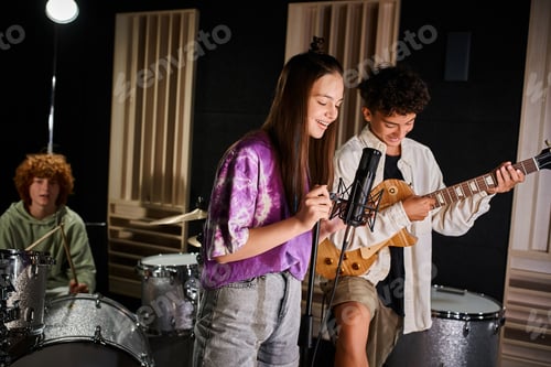 Preview: focus on jolly teenage boy and girl singing and playing guitar next to their blurred drummer