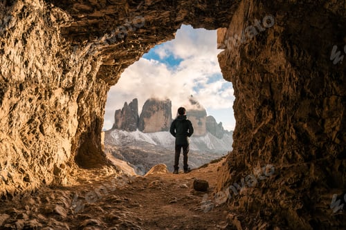 Preview: Male tourist standing at front of rock cave of Grotta delle Tre Cime with mountain peak in Dolomites