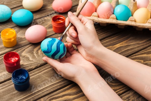 Vista previa: cropped view of woman decorating easter eggs with paintbrush at wooden table