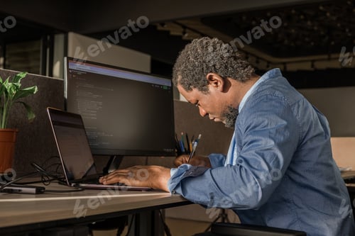 Preview: Black Man Writing Lines of Code On Desktop PC With Two Monitors and a Laptop Aside in Stylish Office