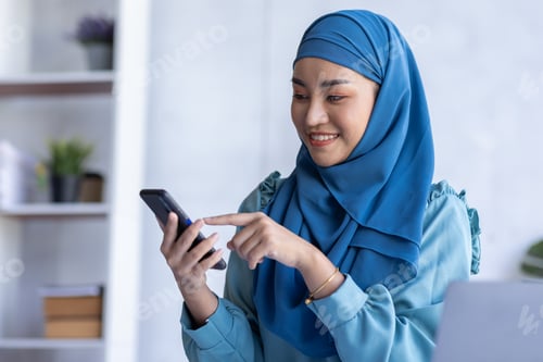 Preview: Smiling Muslim Woman In Hijab Shopping Online With Smartphone While Sitting At Desk In Office, Happy