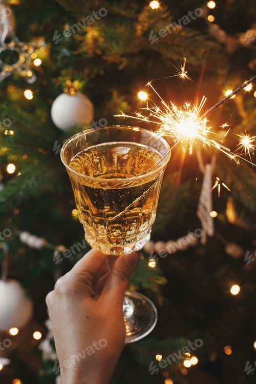Preview: Happy New Year! Hand holding champagne glass with burning firework against christmas tree lights
