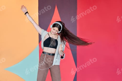 Preview: teenage woman listening to music on smart phone while dancing on a multicoloured wall background