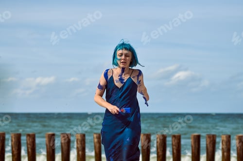 Preview: Artistic blue-haired woman performance artist smeared with blue gouache paints dancing on beach