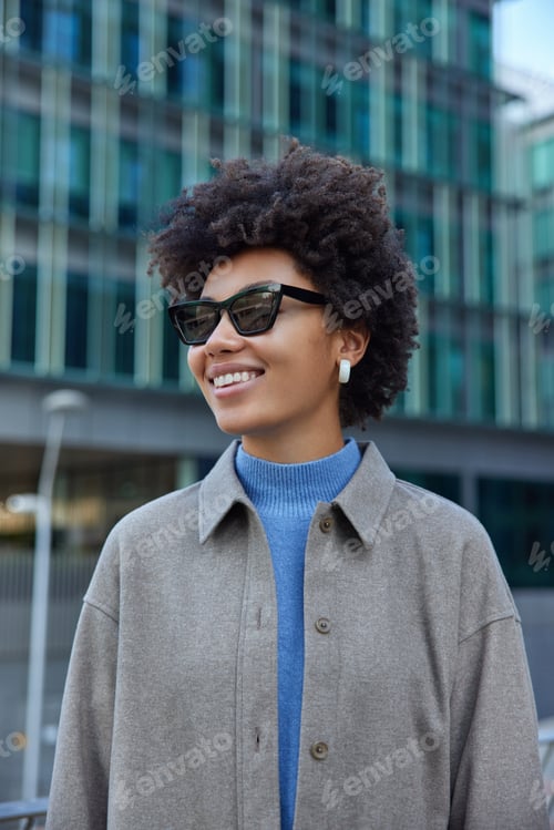 Preview: Stylish female model with toothy smile on face wears sunglasses and grey coat strolls in urban setti