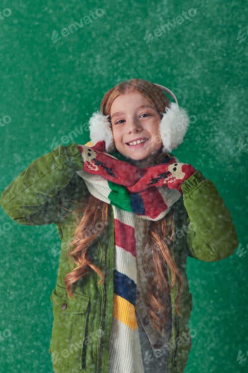 Preview: positive kid in ear muffs, striped scarf and winter attire standing under falling snow on turquoise