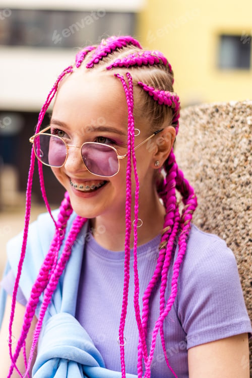 Preview: Smiling caucasian teenage girl with pink braids and with braces on her teeth on a street background.