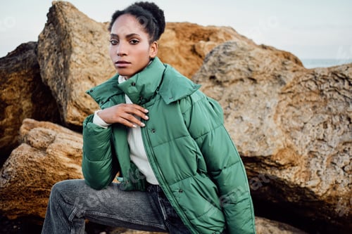 Preview: Gorgeous African American girl in stylish down jacket intently looking in camera on rocks by the sea