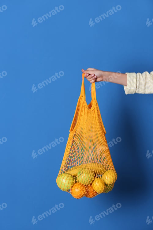 Preview: Woman holding net bag with fruits on blue background, closeup