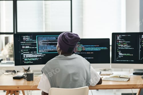 Preview: Back view of young software developer sitting in front of three monitors