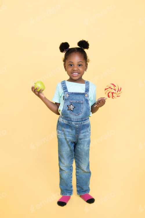Preview: Funny black kid girl, wearing jeans overalls, chooses between a lollipop and a green apple