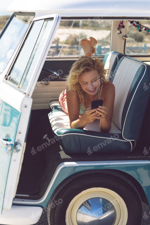 Preview: Front view of happy Caucasian woman using mobile phone while lying on seat in camper van at beach