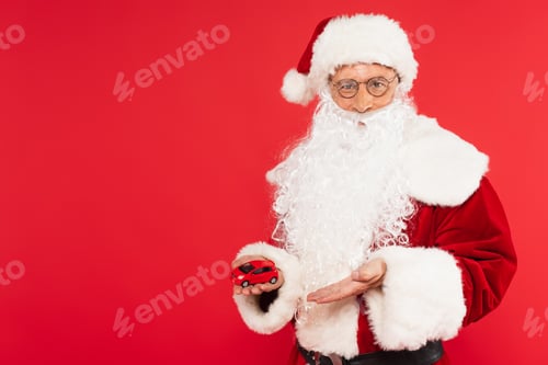 Preview: Santa in hat and eyeglasses pointing at model of car isolated on red