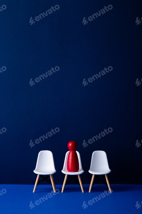 Preview: Red figurine sitting on a chair in a row of empty white chairs.