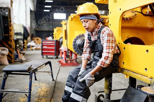 Preview: Young female worker of warehouse in coveralls and hardhat touching hurting knee while sitting by