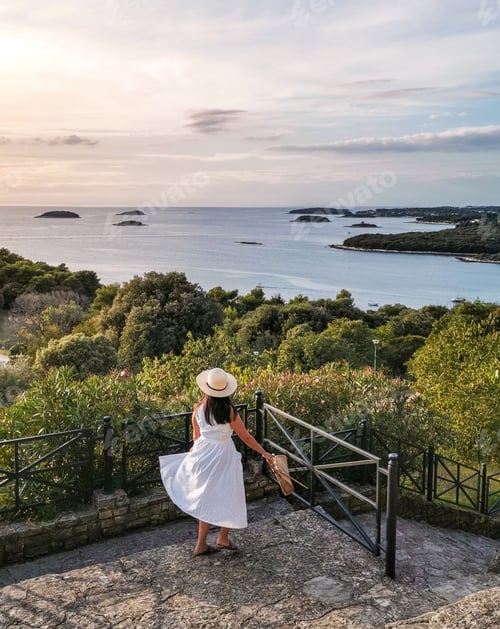 Preview: Young woman in white summer dress and hat on a viewpoint overlooking the sea.