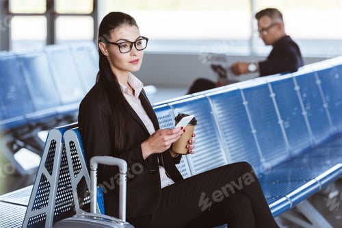 Preview: young businesswoman in glasses sitting with smartphone and coffee to go at airport