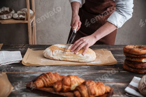 Preview: Cropped image of young man baker cut the bread.