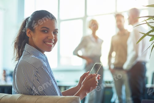 Preview: Young happy mixed race business woman sitting in the modern office, holding her smartphone and