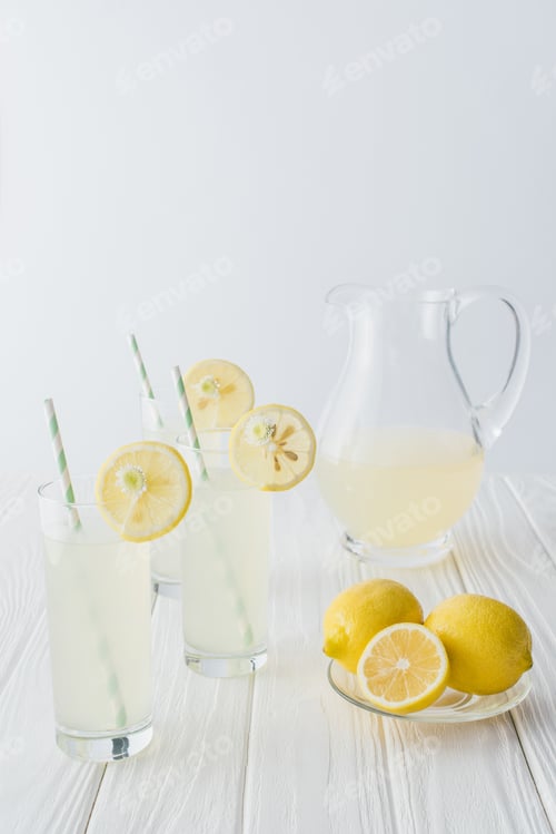 Preview: close up view of lemonade in jug and glasses with straws on white wooden tabletop on grey background