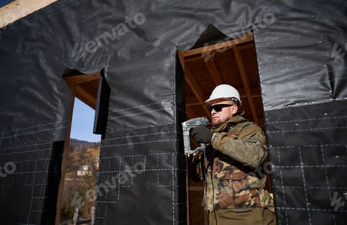 Preview: Male builder installing waterproof membrane on wall of future wooden frame house.