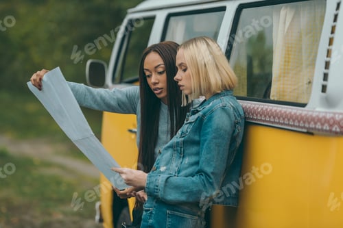 Preview: multiethnic girls looking at map and standing at retro minivan
