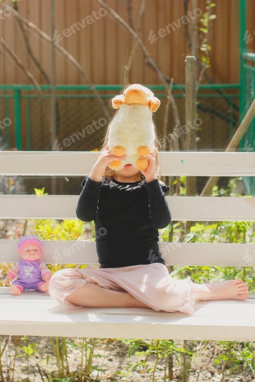 Preview: Sweet little girl hiding behind her toy sheep and sitting in the white bench in the yard