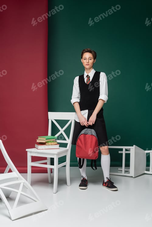Preview: full length of stylish young student in eyeglasses standing with backpack around chairs and books on
