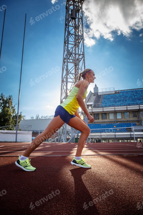 Preview: Sportswoman wearing t-shirt and shorts standing on a red running track in athletics stadium.