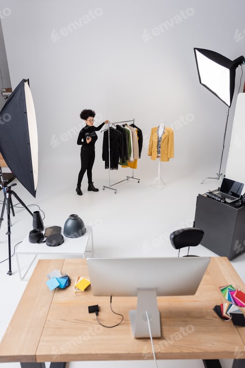 Preview: full length of african american content manager near rail rack with trendy clothing in photo studio