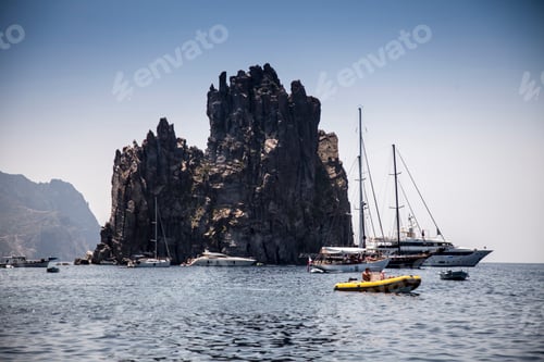 Preview: Sailboats and yachts anchored near a majestic rock formation on a serene sea.